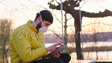 好看的男人，戴着保护面具，在日落时分在公园里，坐在椅子上，他用平板电脑做